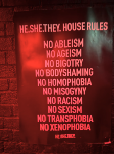 A poster on a brick wall lists house rules against ableism, ageism, bigotry, body shaming, homophobia, LGBTQ discrimination, misogyny, racism, sexism, transphobia, and xenophobia.